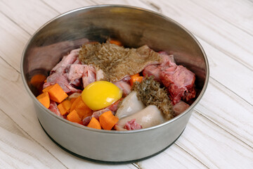 Metal pet bowl with chicken necks, beef meat, paunch, raw egg and carrot pieces. Natural organic daily meal ration preparation for dogs concept. Copy space, white wooden table background.