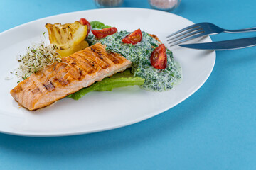 Grilled salmon steak. A piece of red fish on a white plate and a blue background on the table. Fish food with sauce, tomatoes, lemon. Restaurant menu. Grill fried.