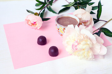 Obraz na płótnie Canvas A Cup of coffee with delicate peonies and two chocolates on a pink background, side view-a place for text-the concept of a pleasant time with friends, loved ones