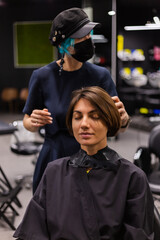 A professional girl hairdresser in face mask makes a client trendy stylish short hair cut. The girl is sitting in stylish beauty salon. Social distance. Protective measures. 