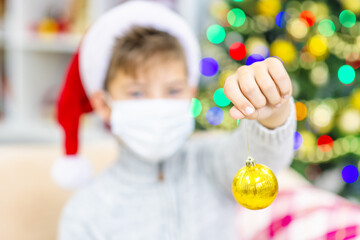 A boy with a medical mask on his face sits at home on the sofa against the background of a Christmas tree in a Santa hat with a Christmas decorations in his hands. Holidays during the coronavirus pand