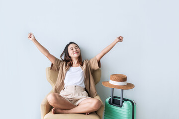 Smiling young Asian traveler girl sitting on couch while raised her hands with happiness gesture on hotel room. Travel alone, summer and holiday concept. Closeup