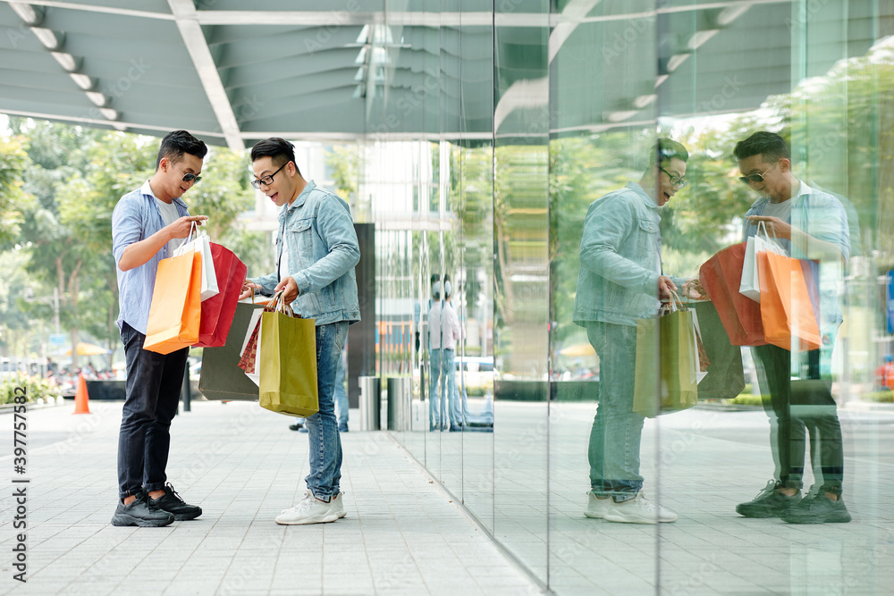 Sticker Stylish young Asian man showing his purchase to excited friend when they are standing in the street