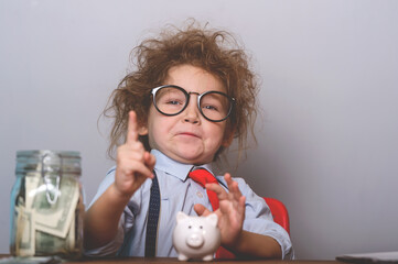 Funny child in glasses and suit pretend to be businessman. Kid playing with money in jar and piggy bank. Education idea dream investment concept.