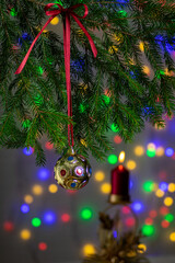 A Christmas tree ball hanging from a red ribbon on a fir branch, against a background of a blurred burning red candle and Christmas tree lights.