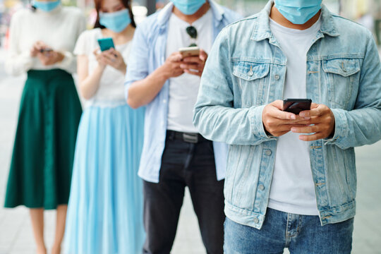 Cropped image of young people in medical masks standing one behind other and texting or checking social media instead of communicating