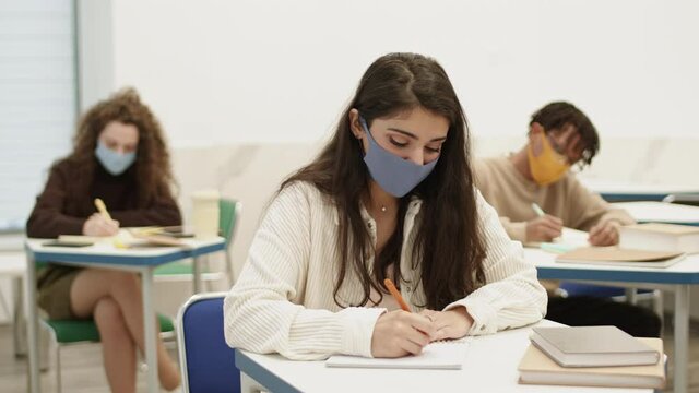 Chest up of Mixed-Race college girl sitting by desk writing in notebook on lecture in classroom. Students learning during pandemic, wearing masks