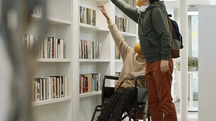 Full shot of disabled African guy in wheelchair looking up, Asian male student taking book off top shelf for him and walking away. People in library wearing face masks - Powered by Adobe