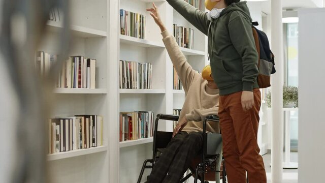 Full shot of disabled African guy in wheelchair looking up, Asian male student taking book off top shelf for him and walking away. People in library wearing face masks