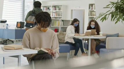 Medium long POV of young African man wearing yellow face mask and glasses, reading book by desk in library, then looking up on camera. Blurred girls sitting at table, unrecognizable student passing by