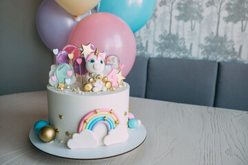 Birthday cake to a girl with a unicorn and balloons..Cake for a girl of gentle pastel tones, decorated with unicorn figures with a bear and rainbow, lollipops, on the background - colorful balls © yulia_rozhno