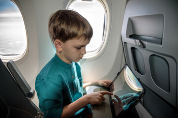 Cute six years old boy, playing on tablen in aircraft on boar, traveling on vacation with parents...