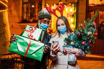 Online shopping for Christmas gifts. Young multiethnic couple in masks choose gifts online. Couple in masks in the middle of the street.