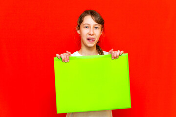 Young self-satisfied teenager girl holding blank paper sheet over red background.