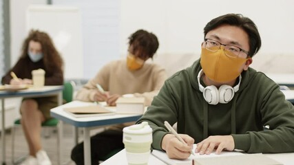 Close-up of Asian male university student wearing mask and glasses, sitting by desk in classroom, taking notes in notebook on lecture. Unrecognizable students learning behind him