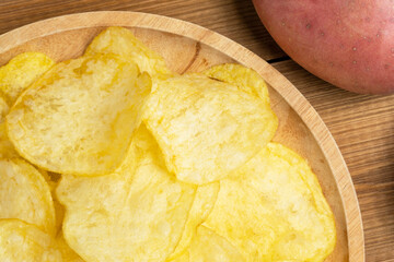Plate of potato chips with raw  potato on wooden table, close up.
