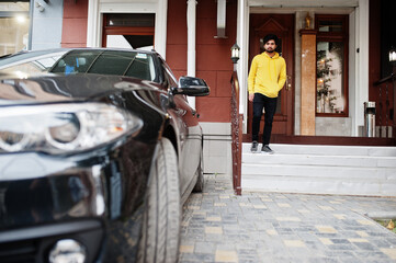 Urban young hipster indian man in a fashionable yellow sweatshirt. Cool south asian guy wear hoodie stand on porch of the house with black business car.