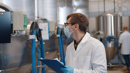 Man in safety mask, gloves and lab coat with clipboard working at craft brewery