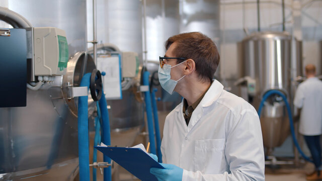 Man in safety mask, gloves and lab coat with clipboard working at craft brewery - Powered by Adobe