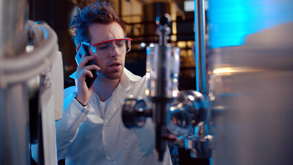 Scientist in lab coat and glass talking on smartphones operating steel tanks on panel