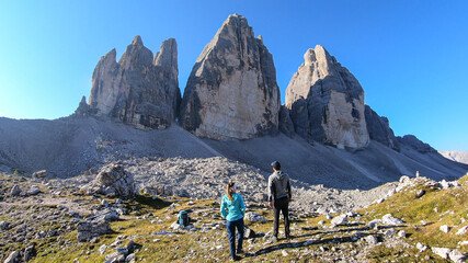 Fototapeta na wymiar A couple enjoying the close up view on the Tre Cime di Lavaredo (Drei Zinnen) in Italian Dolomites. Sharp and high mountain wall. Desolated and raw landscape. Fun and careless moments. Sunny day