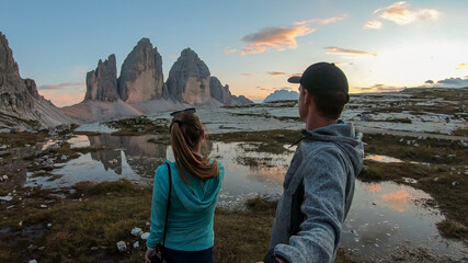 Obraz na płótnie Canvas A couple taking a selfie with the Tre Cime di Lavaredo (Drei Zinnen), mountains in Italian Dolomites. The mountains are reflecting in small paddle. Desolated and raw landscape. Early morning. Daybreak