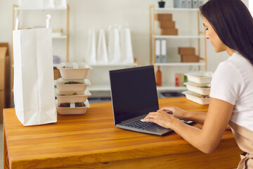 Female food delivery worker turns on laptop to take orders sitting at a table near food containers.