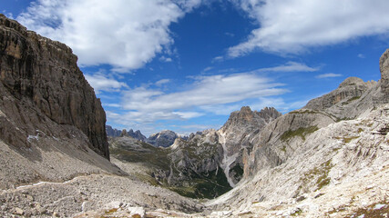 A panoramic view on Dolomites in Italy. There are sharp and steep mountain slopes around. Lots  of lose stones and pebbles. The sky is full of soft clouds. Raw landscape. Serenity and calmness