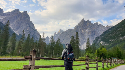 Fototapeta na wymiar A man with a big hiking backpack enjoying the distant view on Italian Dolomites. There is a lush green meadow with a wooden fence around it. Morning fog in the valley. Thick forest on the slopes