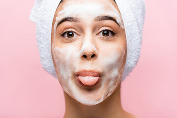 beautiful woman with towel on hair and foam on face showing tongue isolated on pink