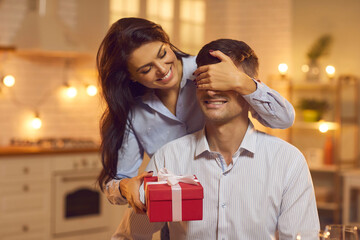 Loving woman stands in the kitchen behind her husband and closes his eyes with her hands giving him.