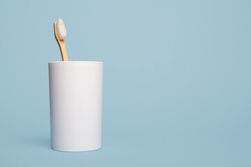 One wooden natural bamboo toothbrush in a white cup on a blue background with copy space and room for text with a left side composition