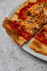 Delicious homemade pizza with tomato, cheese, mushrooms, tuna, vegetables, ham and fresh herbs presented on white plate with light background and selective focus. Copy space.