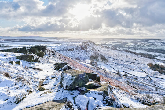 Low winter sun flares through dark cloud over a  snow covered Derbyshire winter landscape