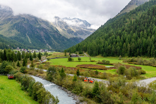 Vacationers stop with their vintage vehicle on the banks of the river that runs through the village of Solda, Ato Adige, Italy, with the mountains covered in low clouds in the background - Powered by Adobe