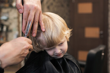 Naklejka premium A little boy is trimmed in the barbershop. Bright emotions on the face, facial expression, the process of cutting hair.