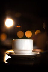 A white ceramic cup on a saucer stands on the table against a background of blurry bokeh-style garland lights. Soft focus. Early or late break for tea and coffee. Festive atmosphere. Vertical content