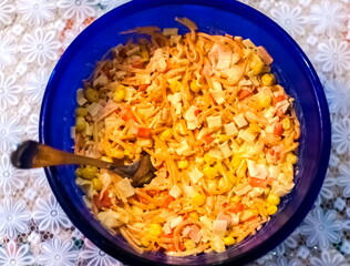 Salad with crab sticks, corn, and Korean carrot with mayonnaise in a blue bowl and a steel spoon closeup on a background of a white tablecloth