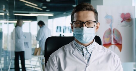 Close up portrait of handsome young male doctor in glasses sitting in hospital office at work, looking at camera and smiling. Healthcare worker in cabinet. Medical profession concept