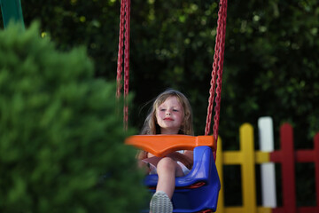 Little girl rides on plastic swing in chains in park on playground in summer.