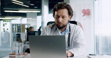Portrait of handsome adult busy Caucasian male doctor sitting at desk in hospital office working and typing on laptop at workplace. Healthcare worker tapping on computer. Job concept