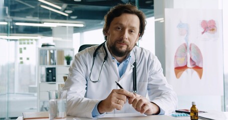 Portrait of beautiful young adult male doctor with stethoscope sitting in clinic at workplace speaking and giving medical advice on consultation. Coronavirus pandemic. Hospital concept