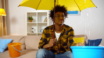 African guy at home under umbrella holding plastic bucket collecting water falling from ceiling.