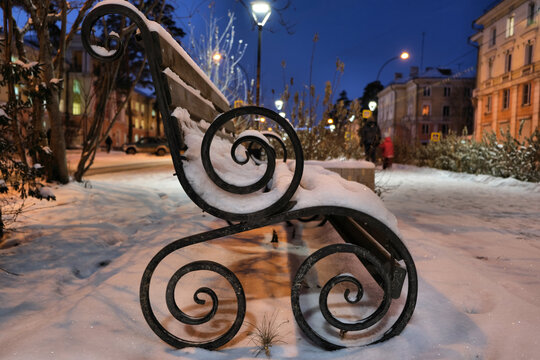 Winter Night Landscape , Bench Covered With Snow Among Frosty Winter Trees And Street Lights. Bench With Black Ornate Cast Iron Legs. Outdoor Furniture