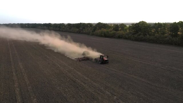 The tractor cultivates the land in the field and applies fertilizer to the soil - the drone orbit shot. Dust billows from the ground behind the cultivator. The concept of the agricultural farming