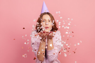 Portrait of carefree cheerful teenage girl in hoodie and party cone blowing heart shaped confetti, enjoying birthday or valentines day, festive mood. Indoor studio shot, isolated on pink background