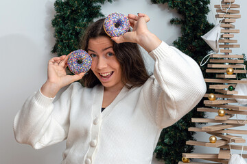 slim brunette in a white knitted jacket holding colorful donuts against her eyes, attractive young...