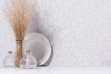 Fototapeta na wymiar Eco nordic home decor with white simple ceramic dish, glass bottles, dried herb bouquet in morning light on white table, copy space.