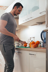 Muscular young man making orange juice in modern kitchen. Active guy preparing healthy meal.