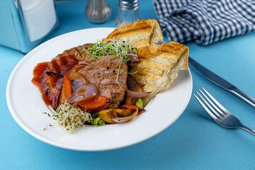 Warm salad with beef, vegetables, croutons and microgreen in a white plate on the restaurant table. Meat food in a bowl. Chop, medallions, cutlets. Blue background, close-up.
